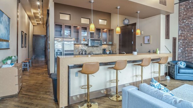 Una cocina moderna con una isla central, taburetes de bar, iluminación colgante y un reloj visible en la pared.