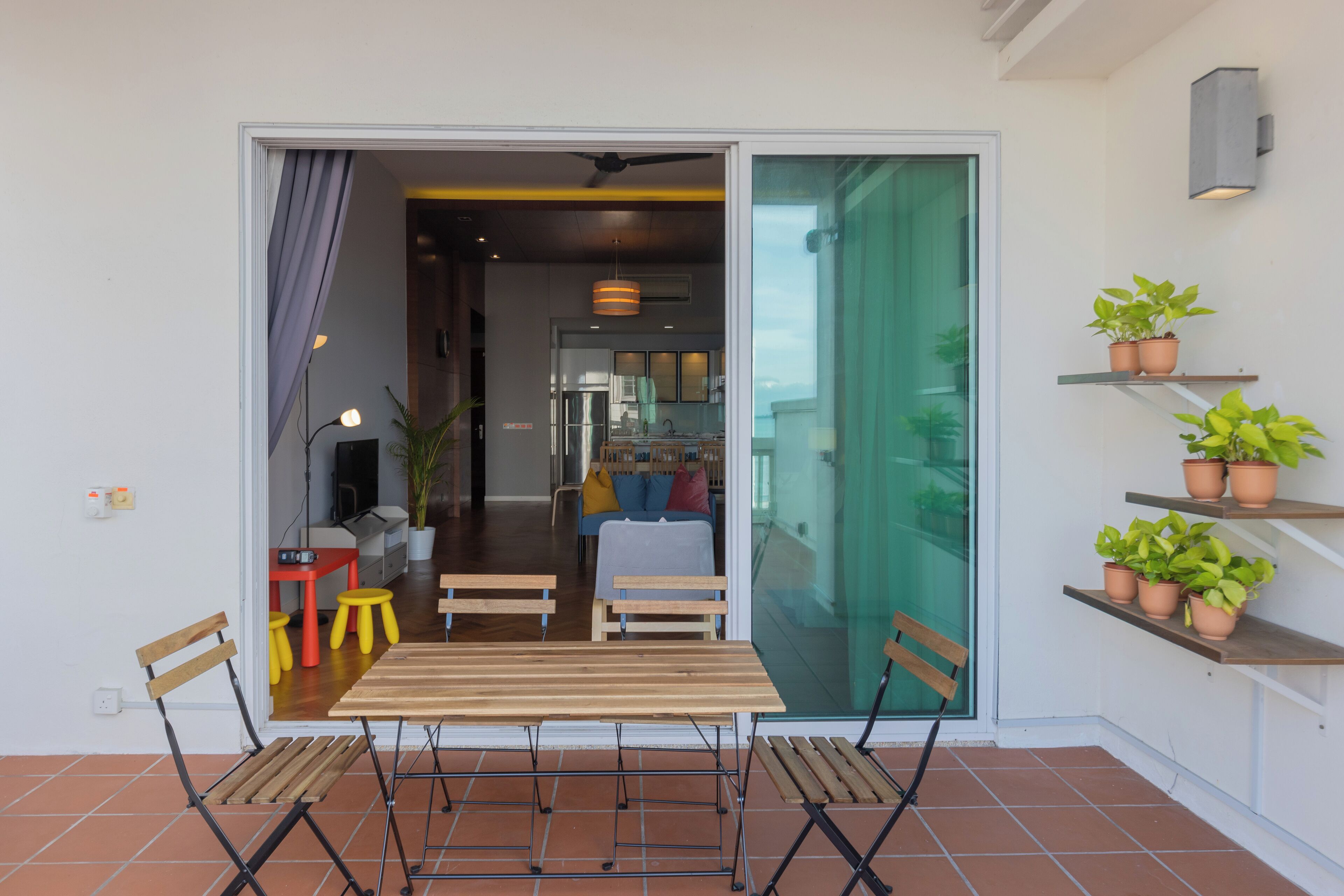 Une porte-fenêtre coulissante donne sur un coin repas avec une table et des chaises en bois, une cuisine à l’arrière-plan et des plantes en pot sur une étagère.