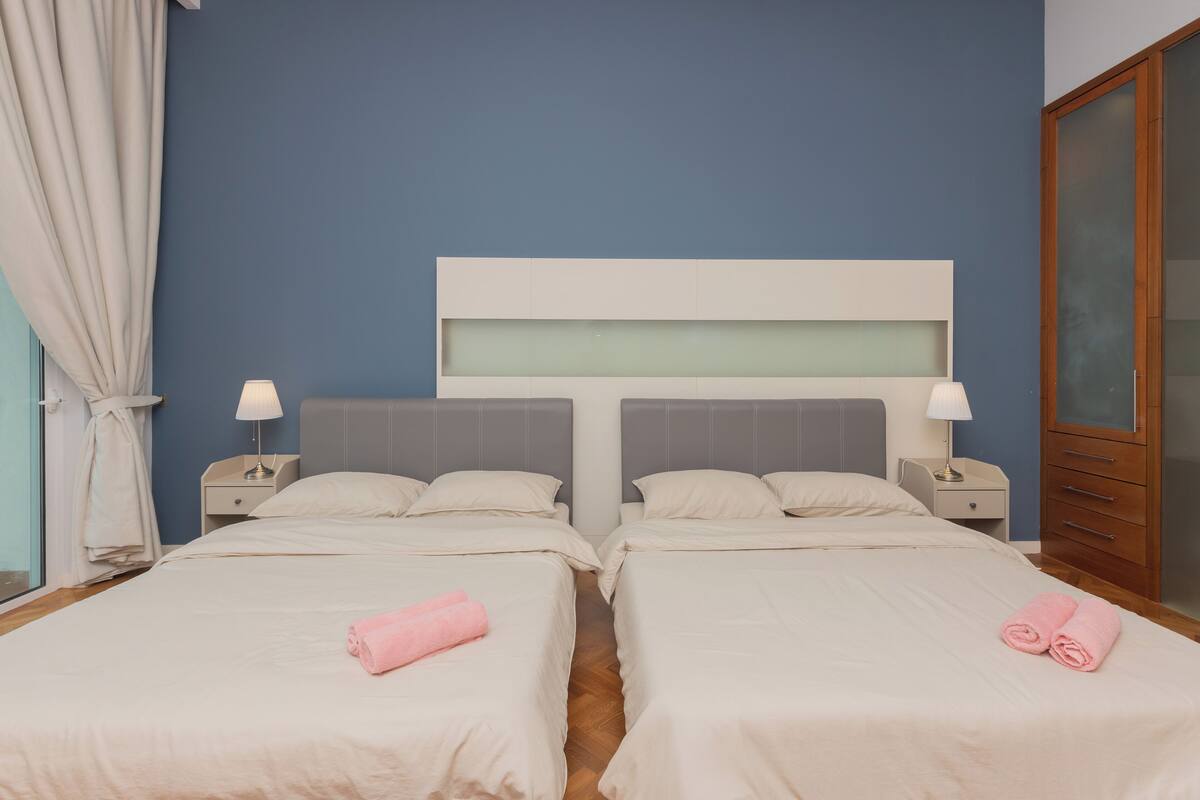 Habitación doble con dos camas, un ventanal grande con cortinas, mesitas de noche con lámparas y un armario.
