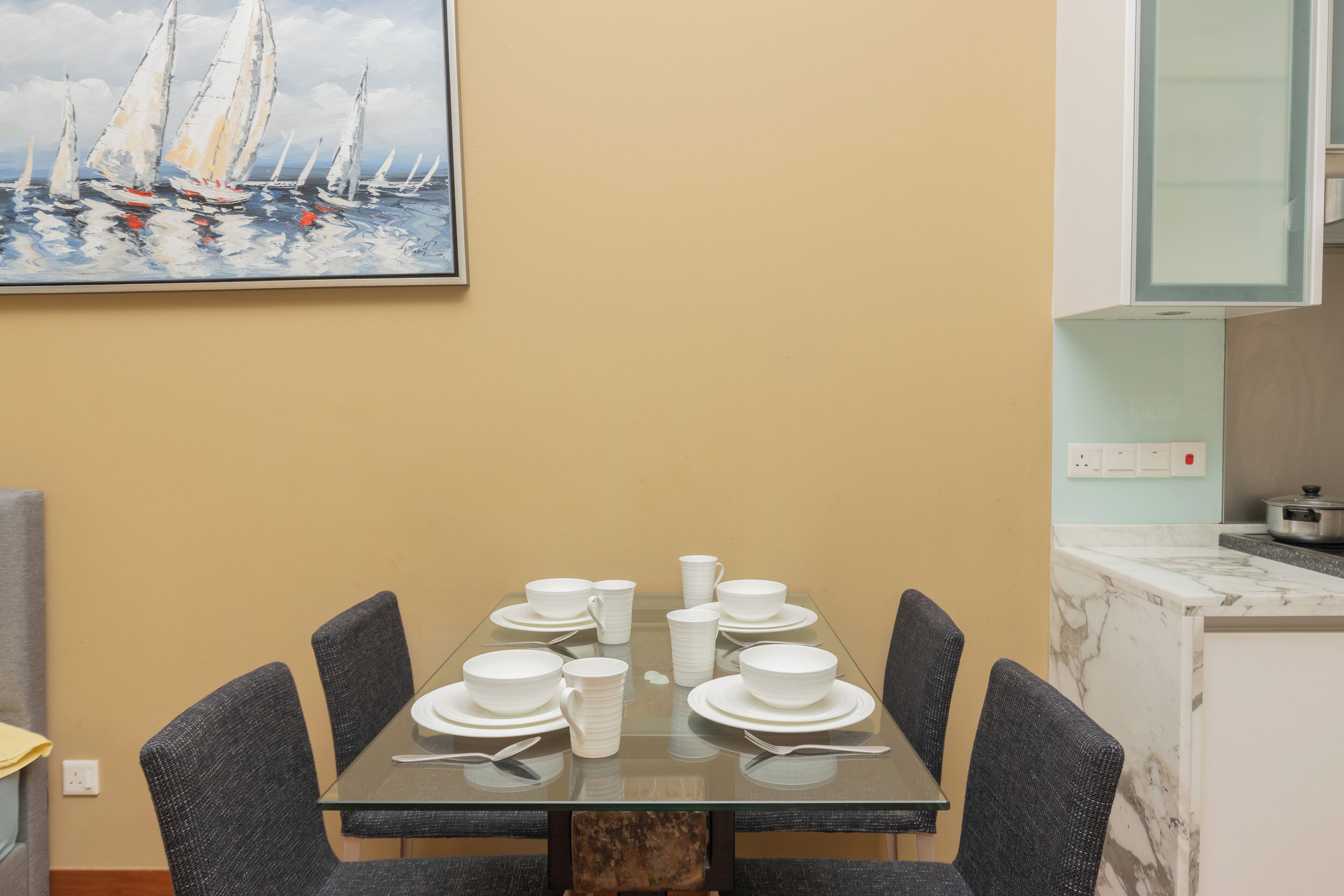 A dining table set with white dishes and cups, surrounded by dark chairs, in a room with a beige wall and a painting of sailboats.