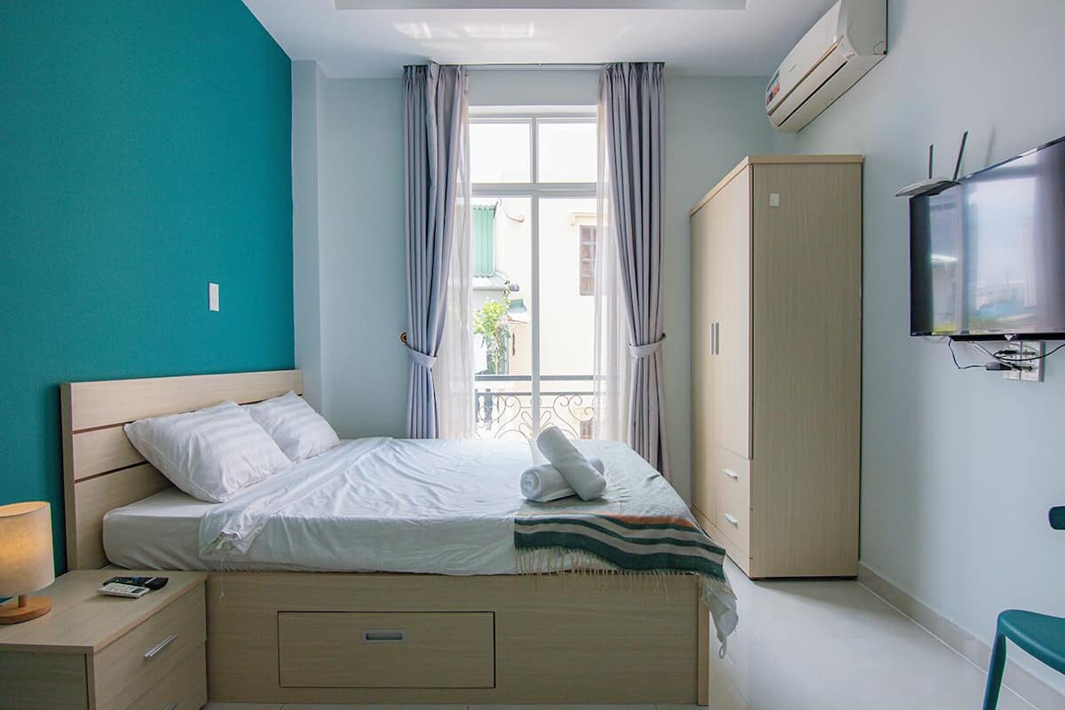 A neatly made bed with a white comforter and pillows, a wooden nightstand with a lamp and remote, a wardrobe, and a television on the wall.