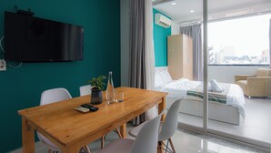 A hotel room with a wooden dining table, chairs, a TV mounted on the wall, a bed with a view of the city, and a balcony with a chair. - Lumina Home (Ho Chi Minh City)