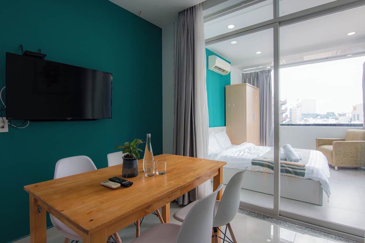 A hotel room with a wooden dining table, chairs, a TV mounted on the wall, a bed with a view of the city, and a balcony with a chair.