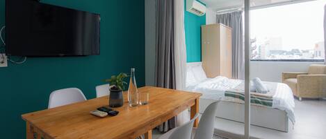 A hotel room with a wooden dining table, chairs, a TV mounted on the wall, a bed with a view of the city, and a balcony with a chair.