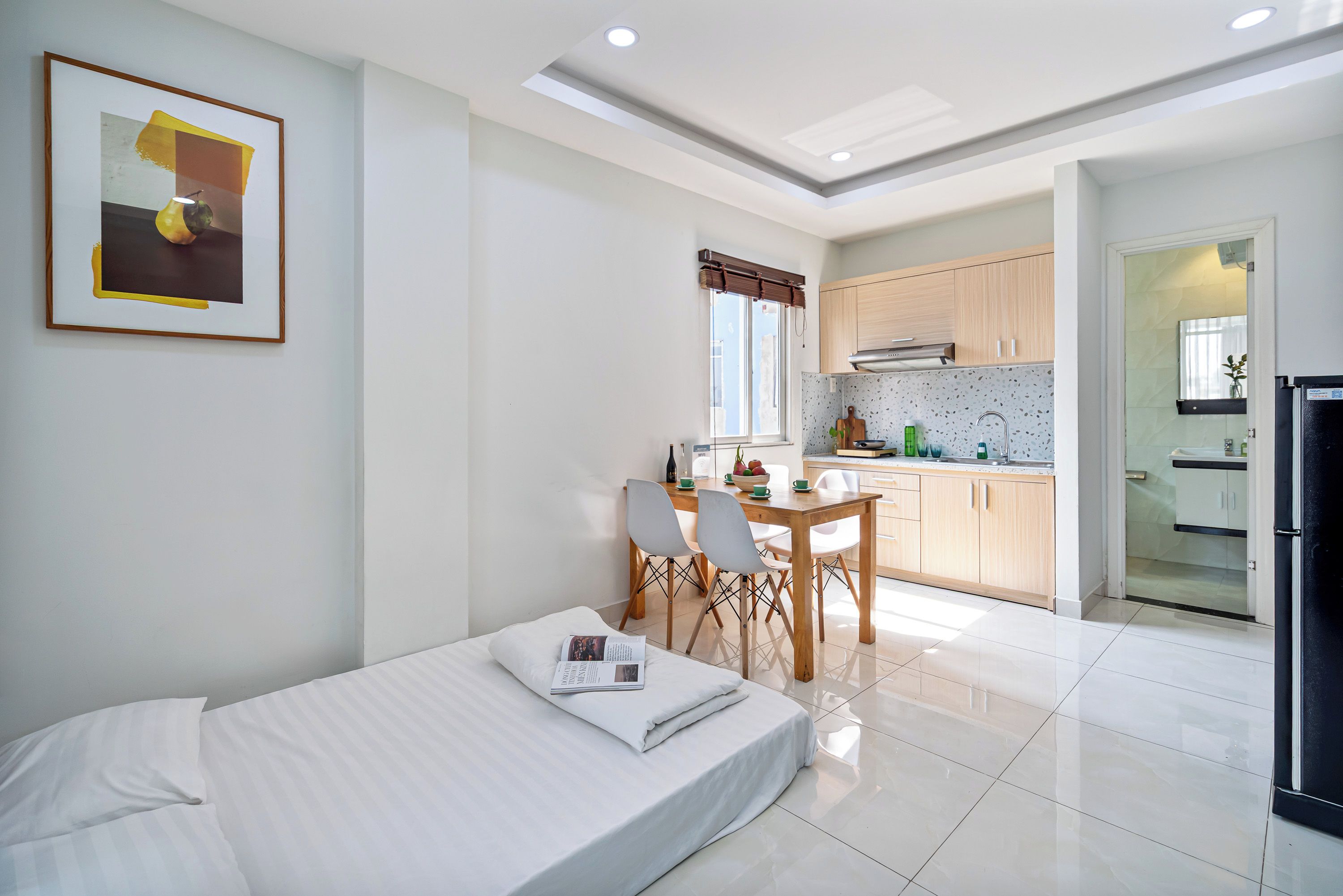 A modern hotel room with a bed, a dining table with chairs, a kitchen area, and a bathroom.
