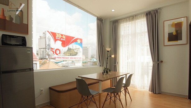 A modern living room with a wooden dining table, two chairs, a bench, a refrigerator, and a large window with curtains. - Lumina Home (Ho Chi Minh City)