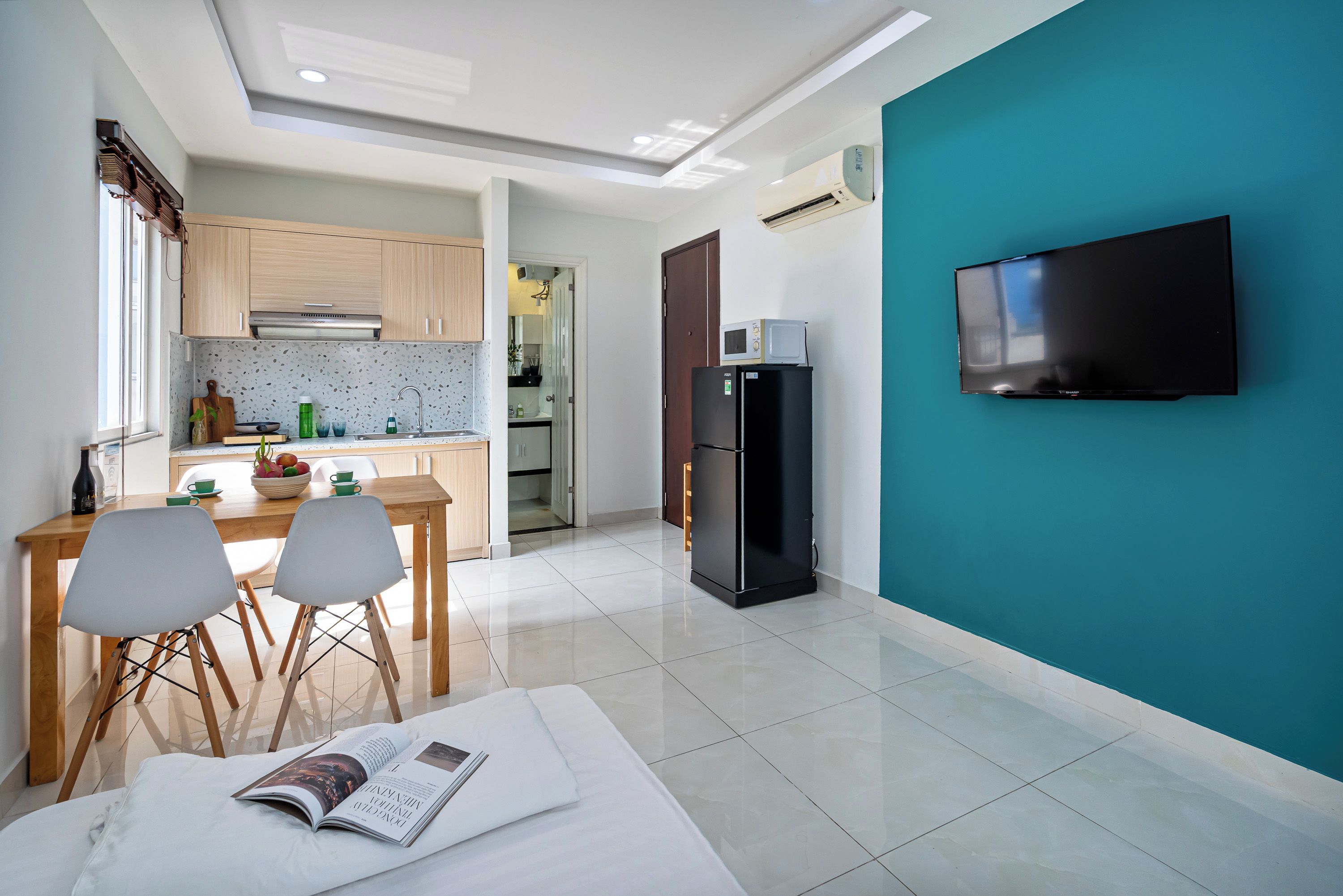 A modern kitchen with wooden cabinets, a dining area with chairs, a black refrigerator, and a flat-screen TV mounted on a teal wall.