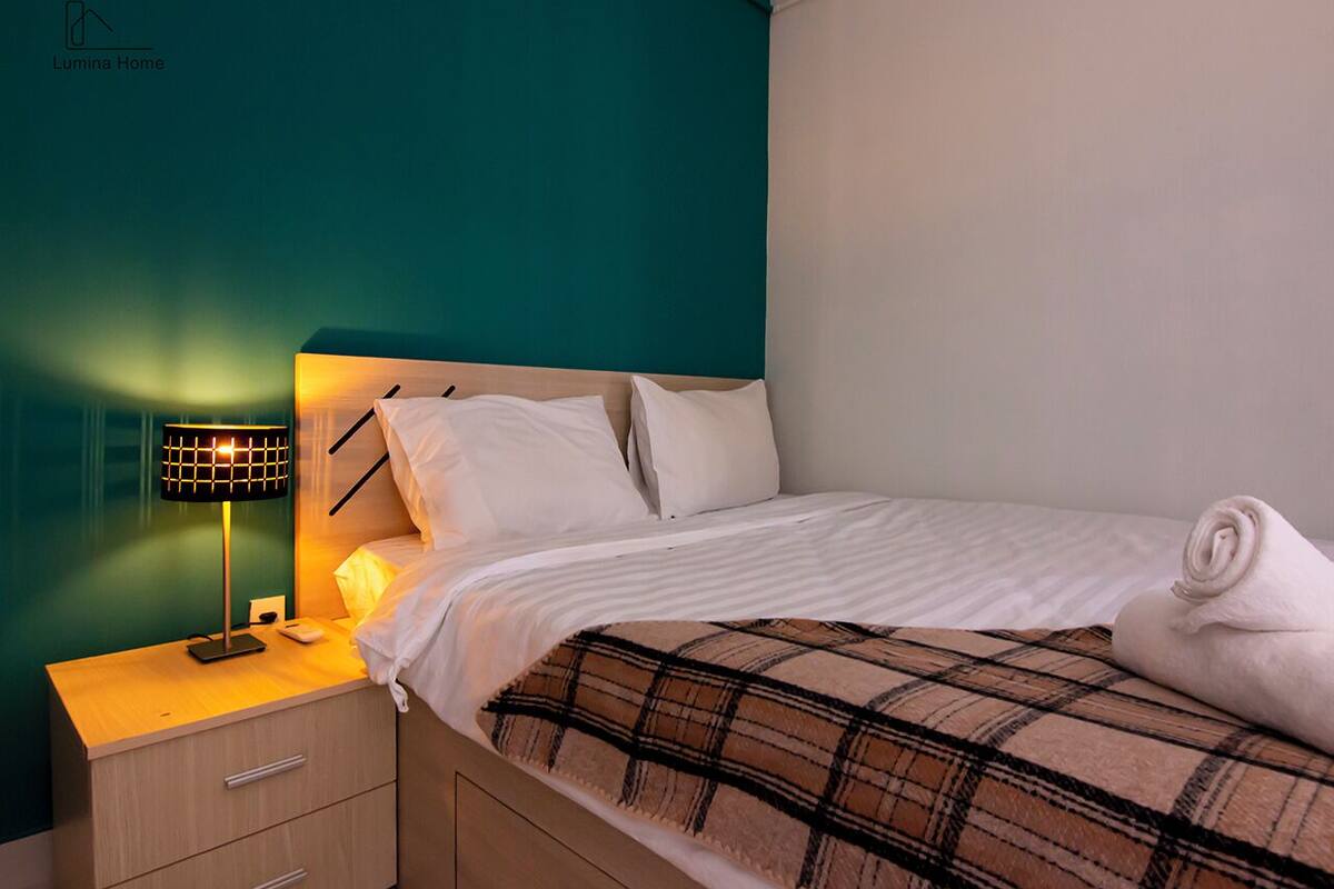 A neatly made bed with white linens and a plaid blanket, a bedside lamp, and a small wooden nightstand with a drawer.