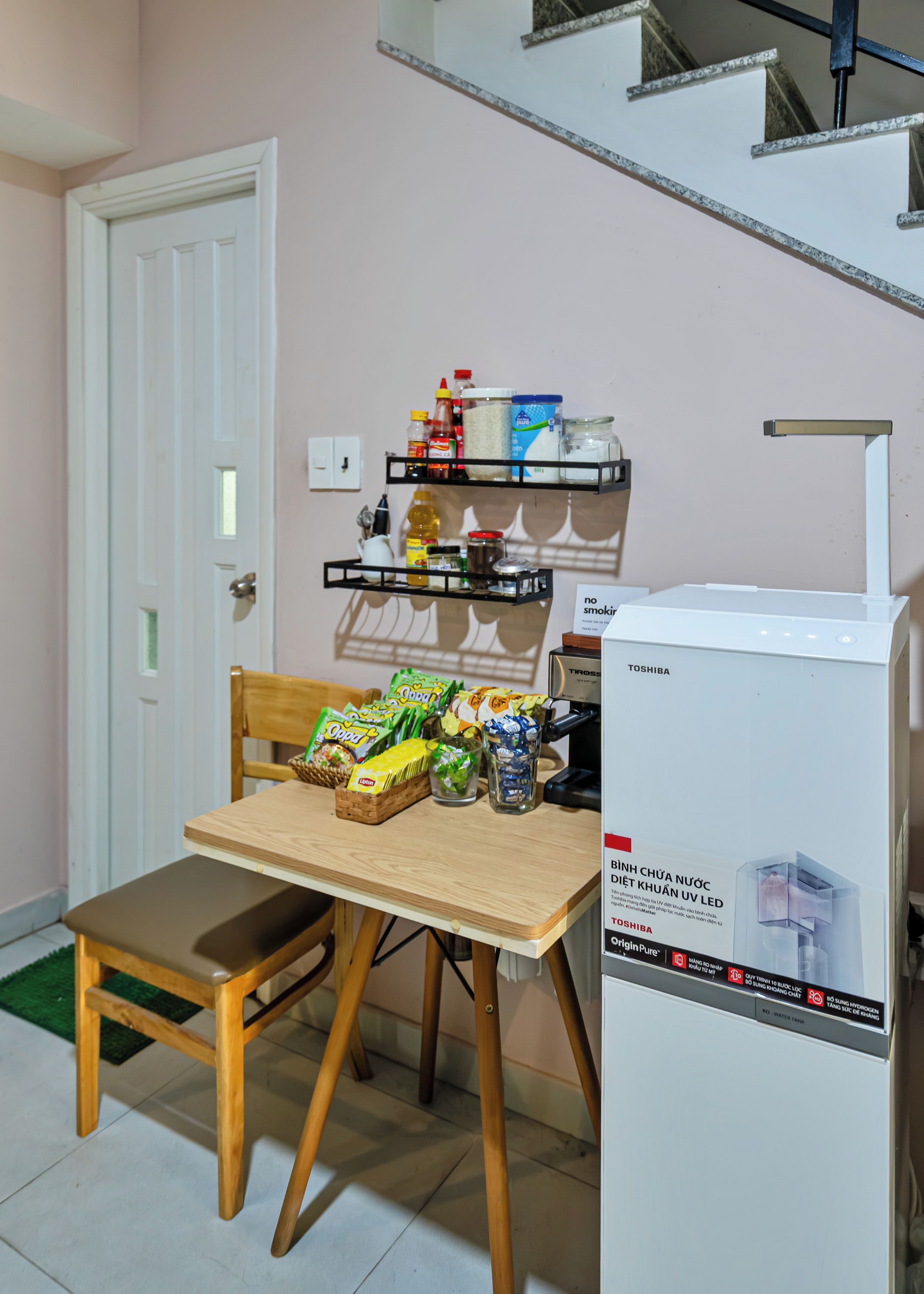 A kitchenette with a refrigerator, a wooden table with chairs, and shelves above holding various food items and condiments.