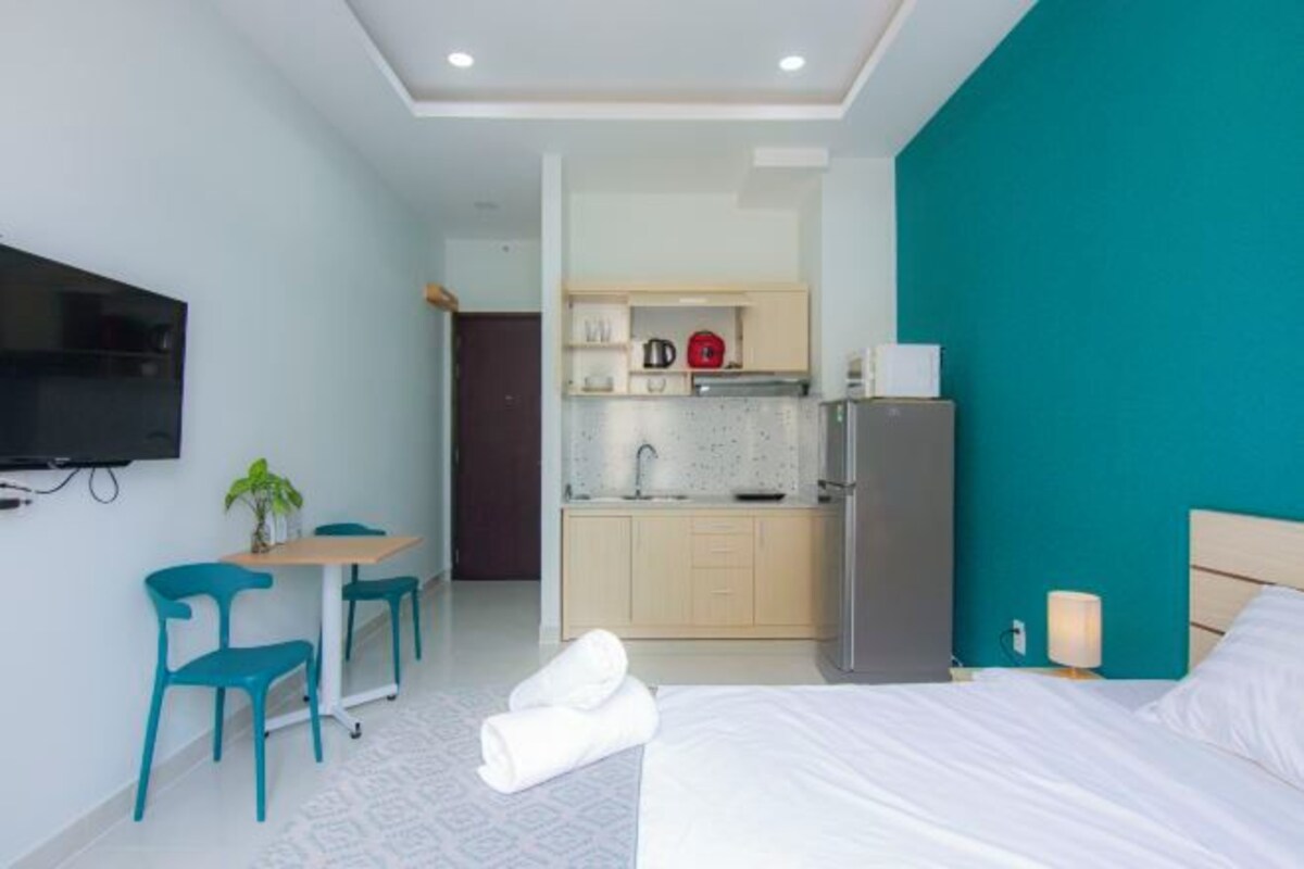 Una habitación de hotel con cama, escritorio con sillas, kitchenette y televisión instalada en la pared.