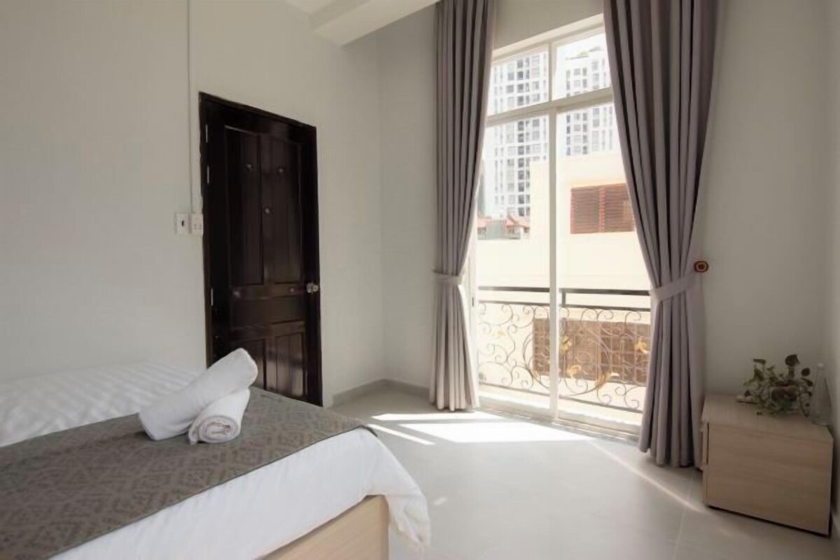 A bedroom with a bed, a window with curtains, a door, and a nightstand with a plant.