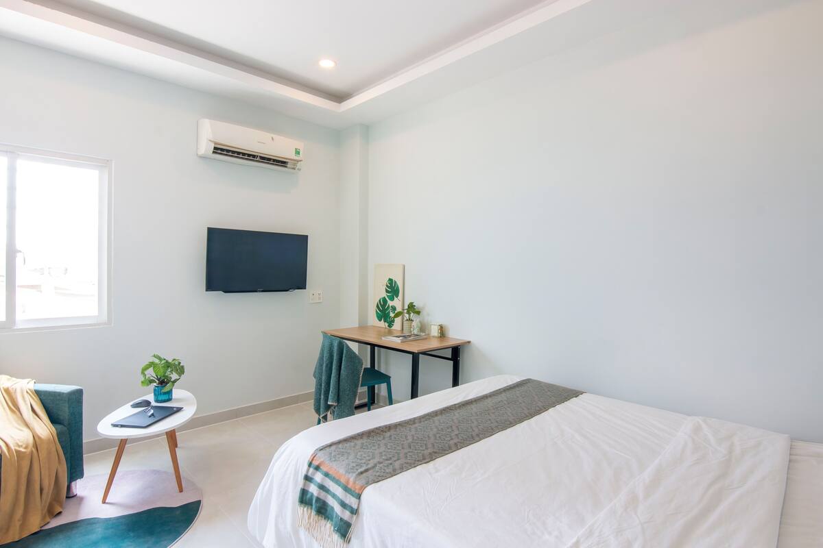 A hotel room with a bed, a desk, a chair, a small table with a plant, and a TV mounted on the wall.