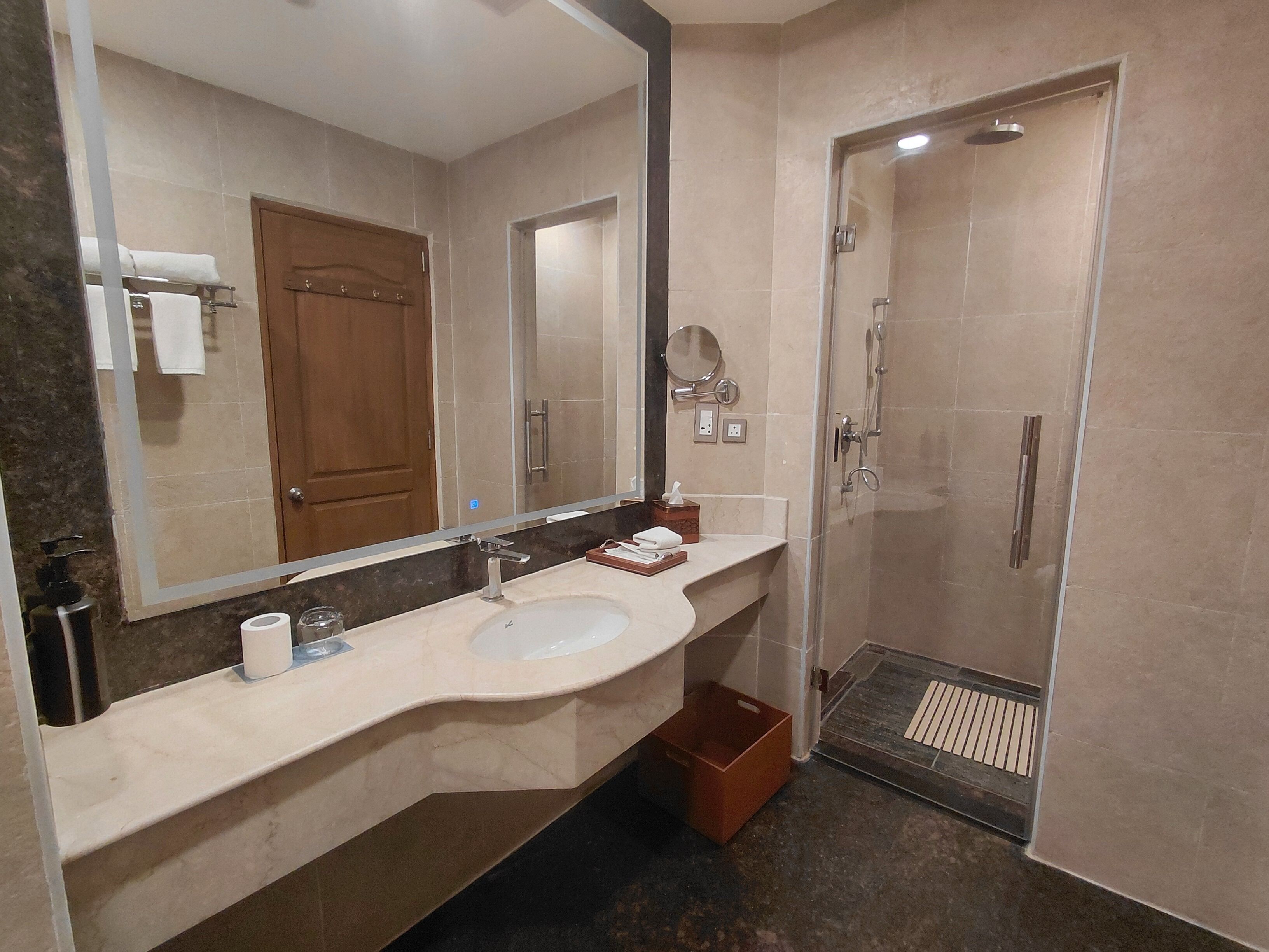 A bathroom with a sink, mirror, towel rack, shower, and a trash bin.