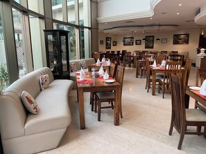 A spacious dining area with multiple wooden tables and chairs, a sofa with floral cushions, and large windows offering a view of the outdoors. - The BellaVista Hotel Chandigarh Panchkula (Panchkula)