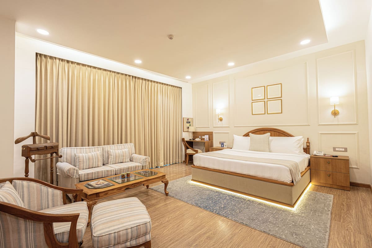 A hotel room with a large bed, a sofa, a chair, a wooden nightstand, and a desk with a lamp.