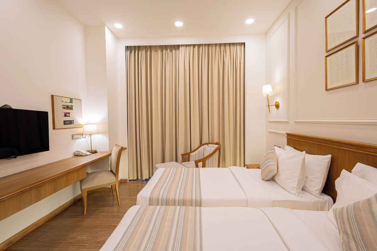 A hotel room with two beds, a desk, a chair, a television, and a large window with curtains.