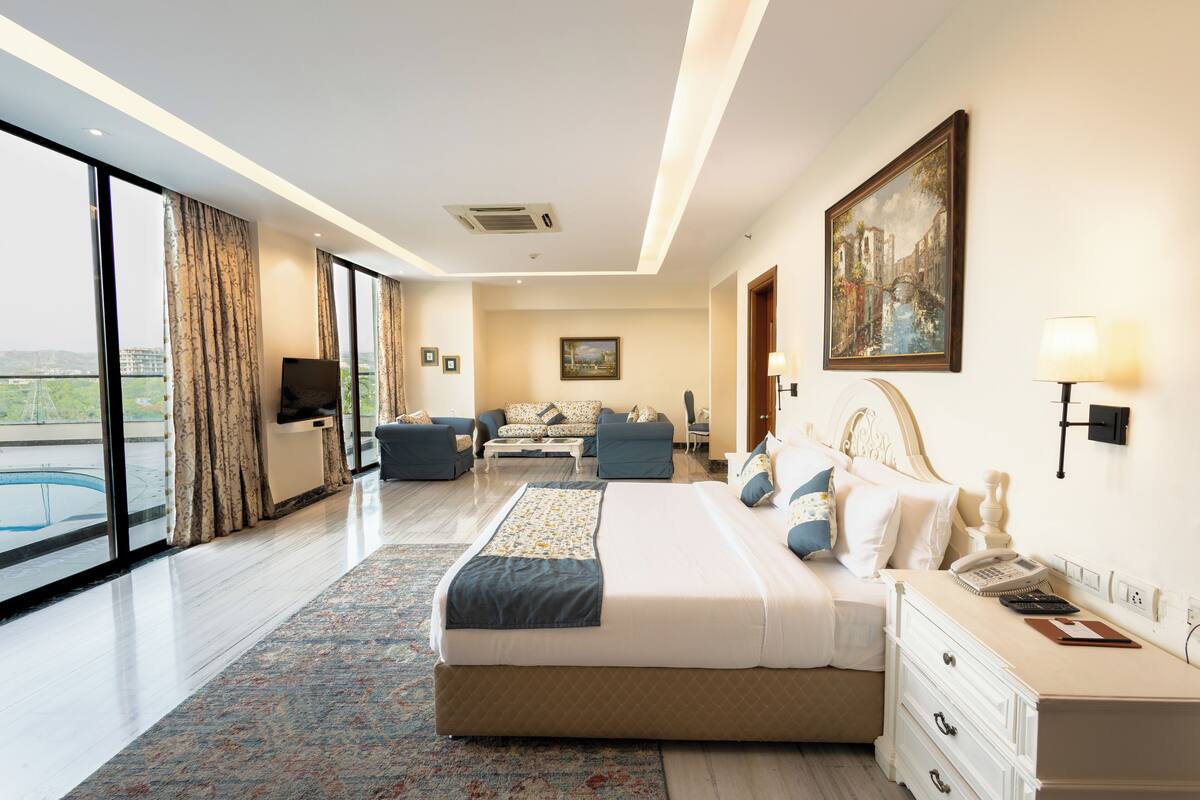 A modern hotel room with a large bed, a desk, a chair, a sofa, and a view of the city.