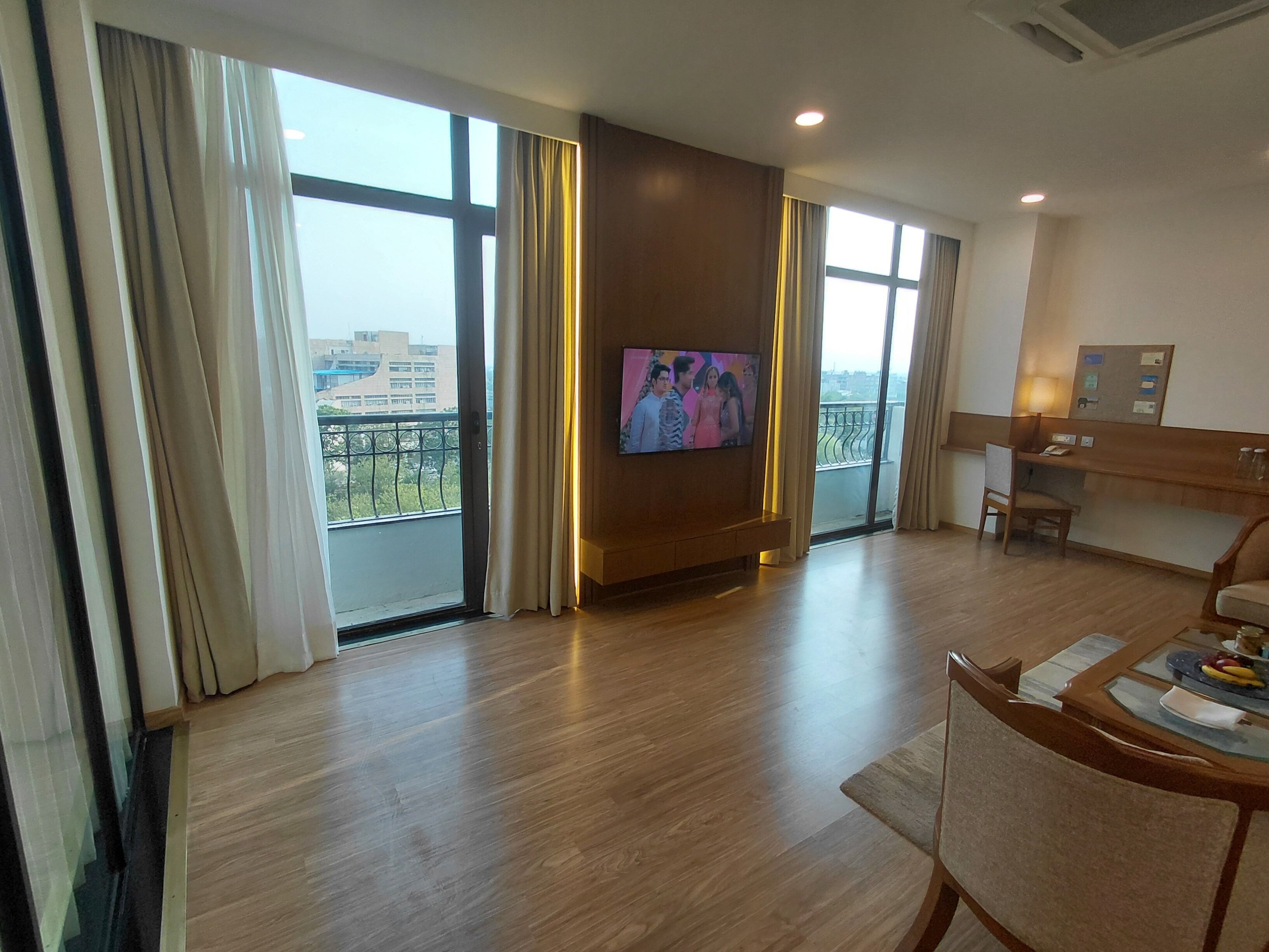 A hotel room with a large TV, a balcony with a view, a desk with a chair, and a wooden floor.