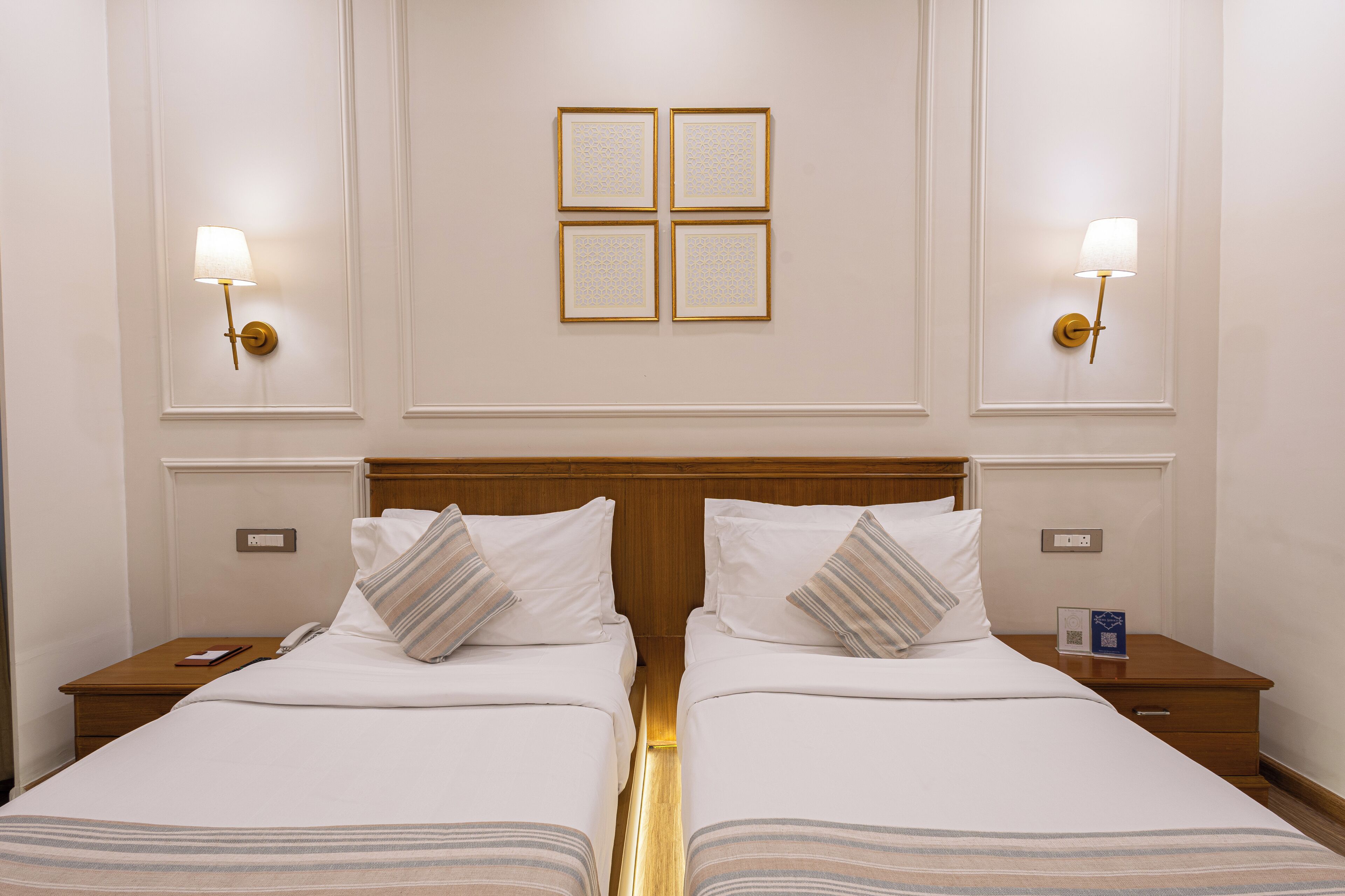 A hotel room with two beds, nightstands, a wooden headboard, wall sconces, and a framed artwork above the beds.