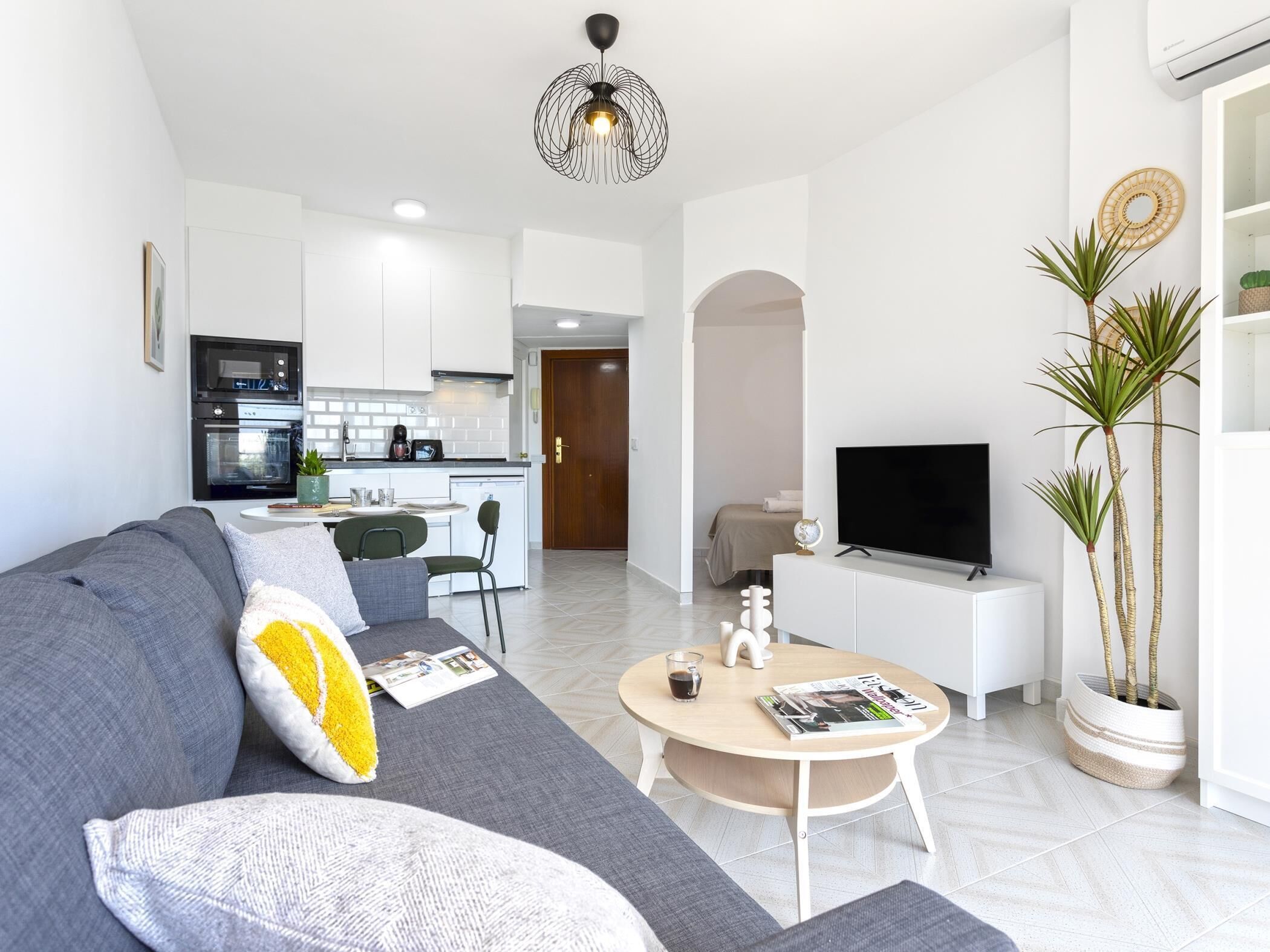 Una sala moderna con un sofá gris, una mesa de centro, un televisor y una cocina al fondo.
