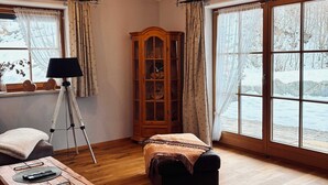 Une pièce chaleureuse avec un plancher en bois, une grande fenêtre donnant sur un paysage enneigé, un meuble en bois et un trépied avec une lampe.