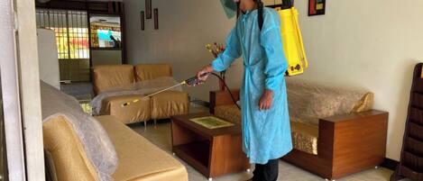 A person in protective gear using a spray device in a living room with sofas and a wooden coffee table.