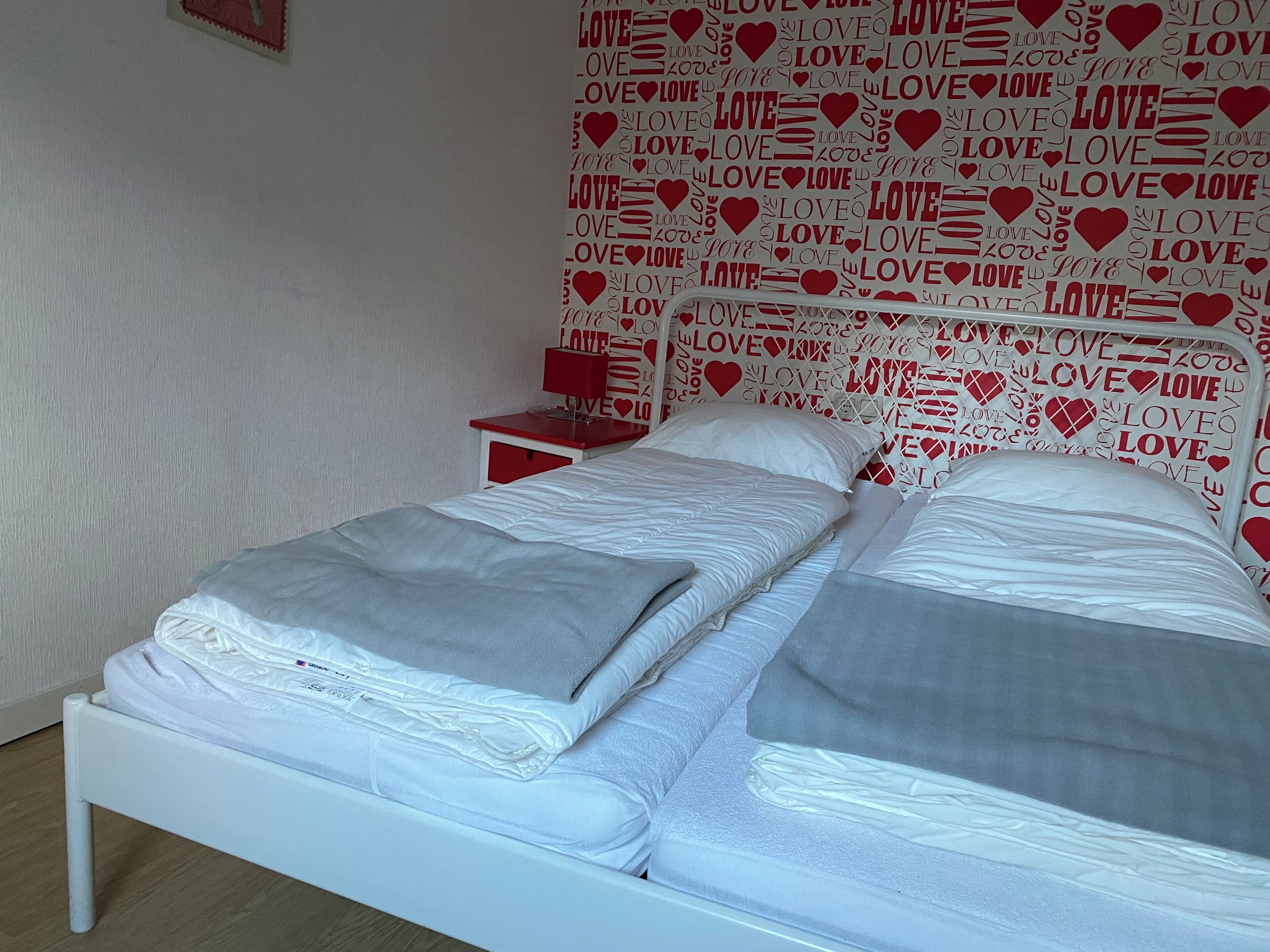 Een bed met witte lakens en kussens, een rode nachtkastje, en een muur met het patroon van het woord "LOVE" en harten.
