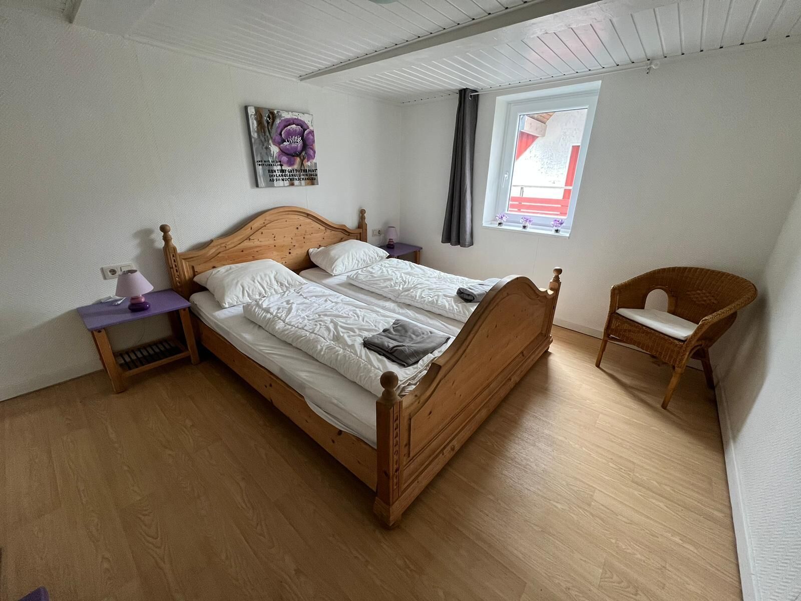 A single wooden bed with white bedding, a small bedside table with a lamp, a wicker chair, and a window with curtains.