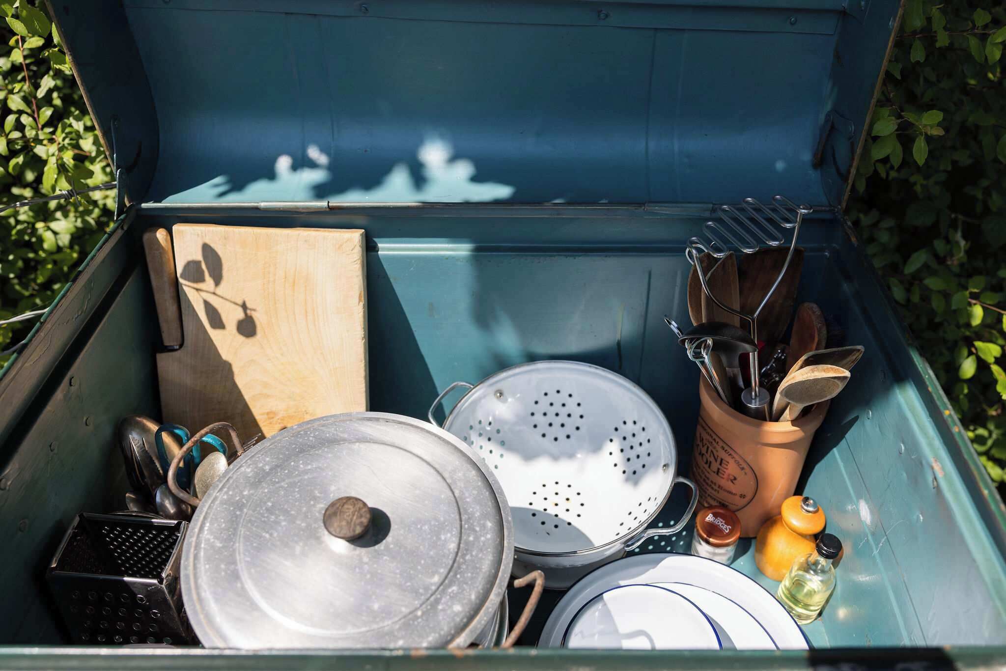 A blue storage container with kitchen utensils, a colander, a wooden box, and a metal pot lid.