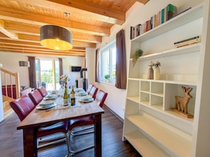 A dining area with a wooden table set for four, a bookshelf with books and decorative items, and a wooden ceiling with exposed beams.