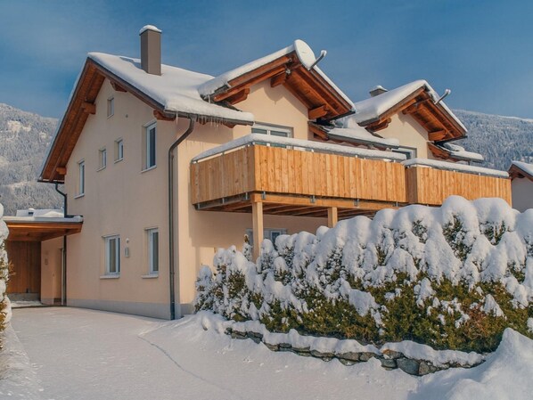 Un chalet de dos pisos con balcón de madera, techo cubierto de nieve y un muro de piedra al frente.