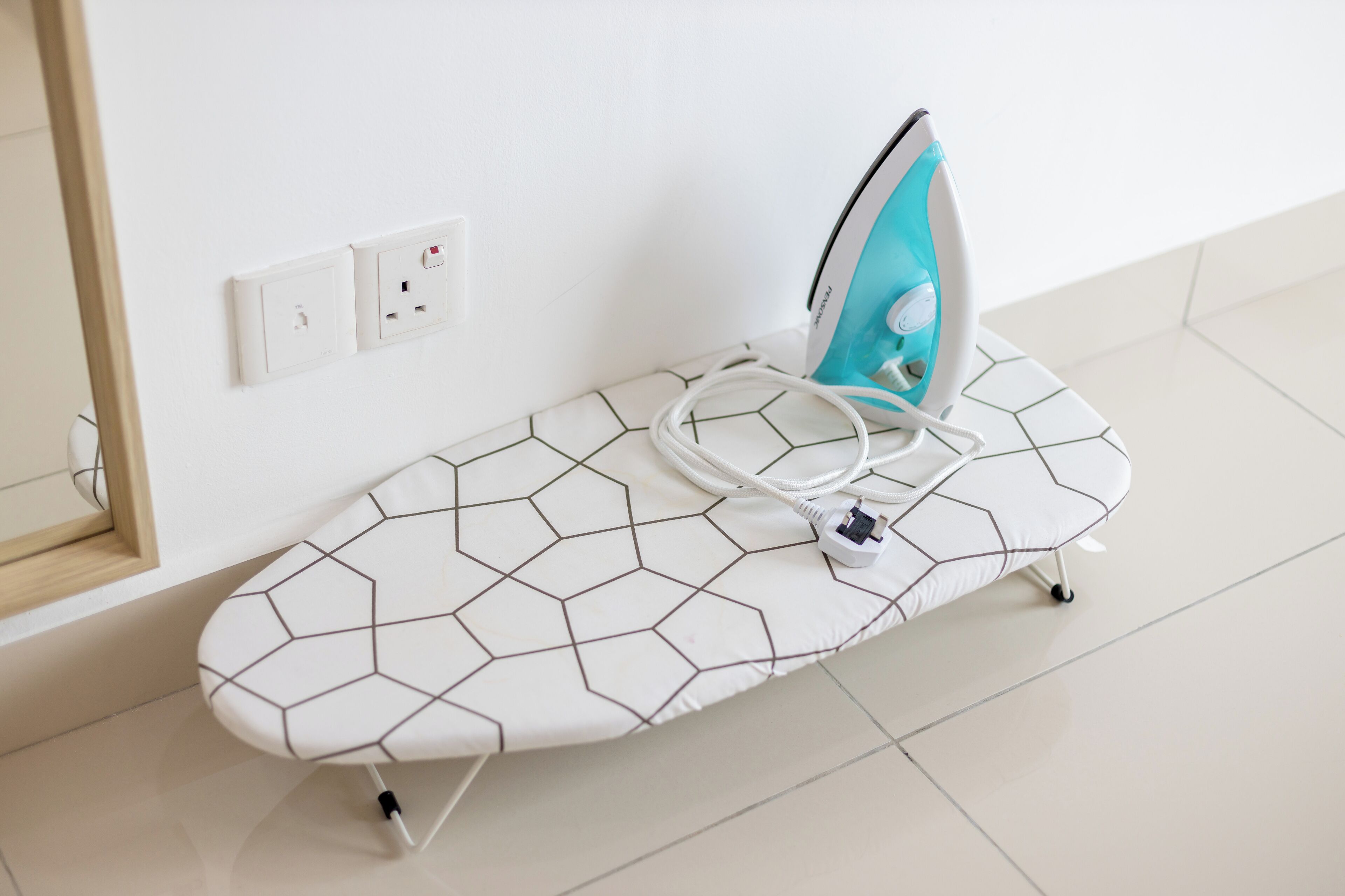 A white ironing board with a geometric pattern, a blue iron, and a power cord plugged into a wall socket.