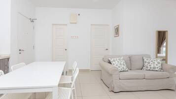 A modern living room with a white sofa, a dining table, and chairs. There are two doors, one with a light switch above it, and a framed picture on the wall.