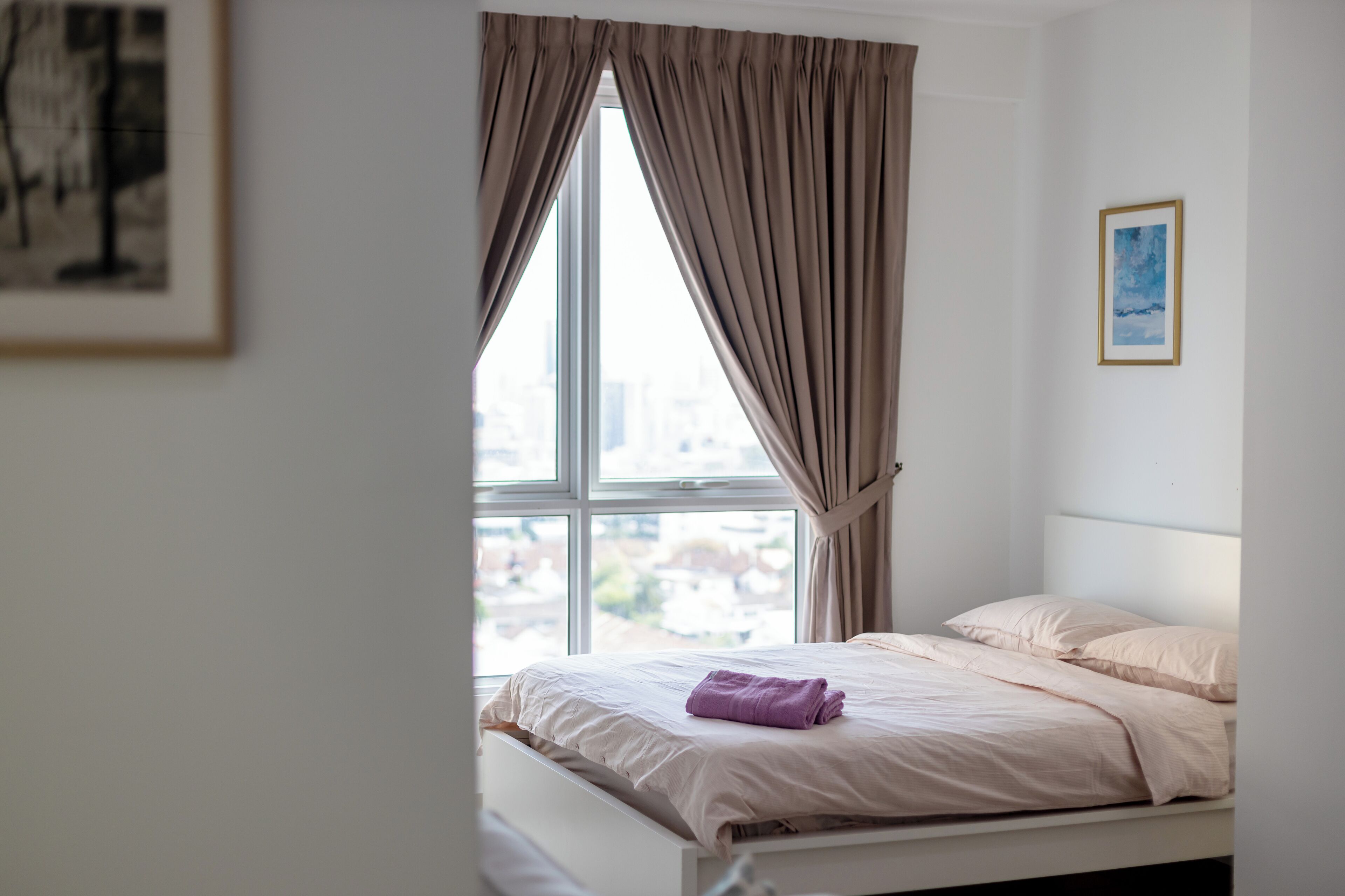 Une chambre à coucher avec un lit, une fenêtre avec des rideaux, un tableau encadré au mur et une vue sur les bâtiments à l’extérieur.