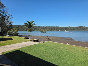 A grassy area with a concrete path leading to a beach by a lake, surrounded by trees and hills.