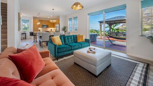 A spacious living room with a teal sofa, a grey ottoman, and a red couch. A kitchen is visible in the background, and a large sliding glass door leads to an outdoor area with a hammock and a view of the ocean.