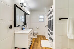 Et moderne badeværelse med en hvid vask, spejl og toilet, trægulv samt bruseforhæng med et geometrisk mønster.