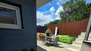 Eine Terrasse mit Tisch und Stühlen, ein Holzregal mit Blumentöpfen und Blick auf einen Garten mit einer Hecke.