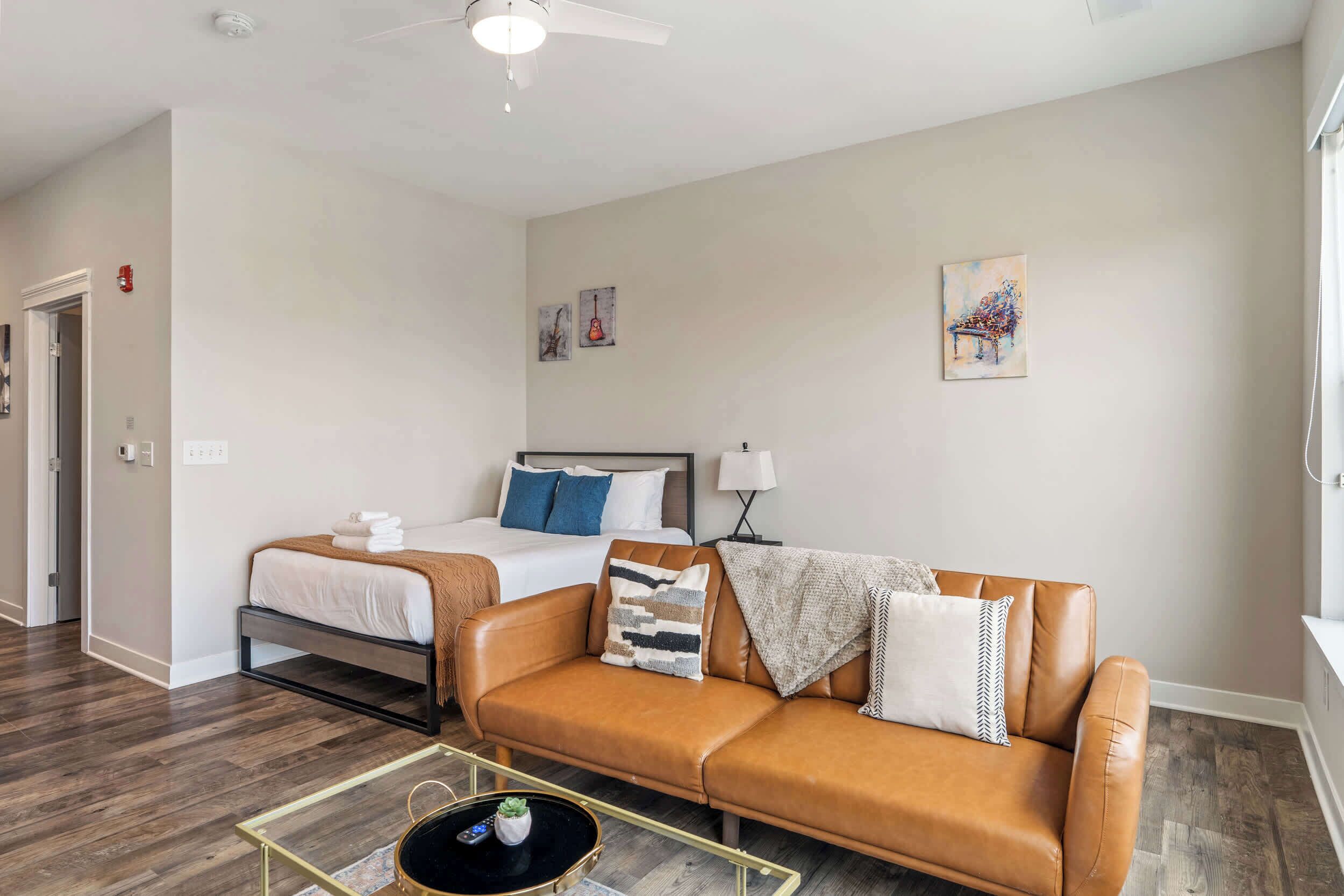 Een moderne hotelkamer met een bruine leren bank, een bed met witte beddengoed, een tafeltje met een afstandsbediening en kunstwerken aan de muren.