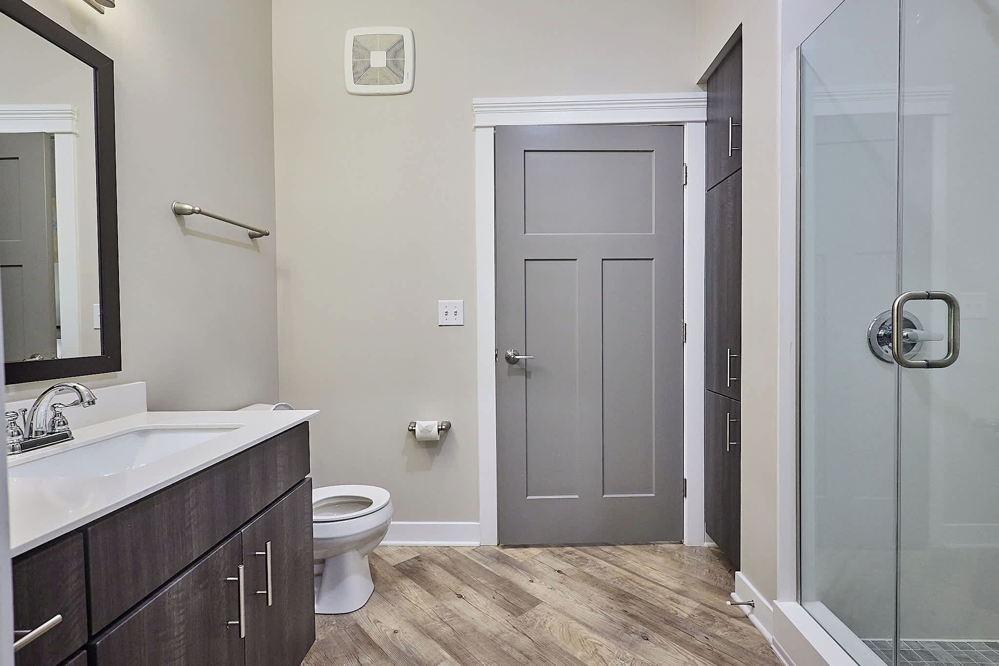 Une salle de bain avec un lavabo blanc, des meubles en bois foncé, des toilettes, une porte et une cabine de douche en verre.