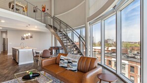 A spacious living area with a high ceiling, a staircase, a kitchen, and large windows offering city views. - CozySuites - Penrose (Indianapolis)