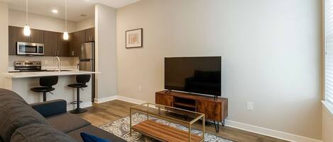 A modern living room with a sofa, a coffee table, a TV, and a kitchen area with a microwave and bar stools.