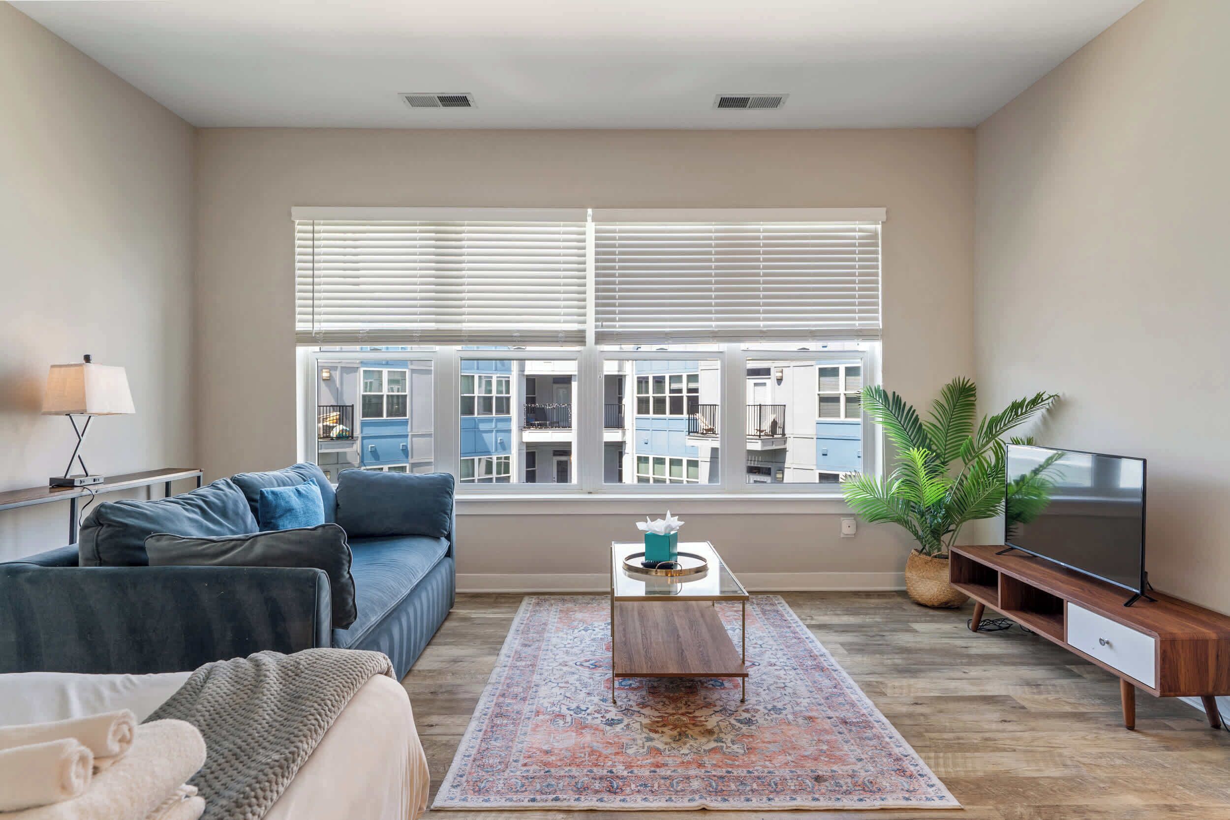 A modern living room with a sofa, coffee table, TV, and large windows with blinds.