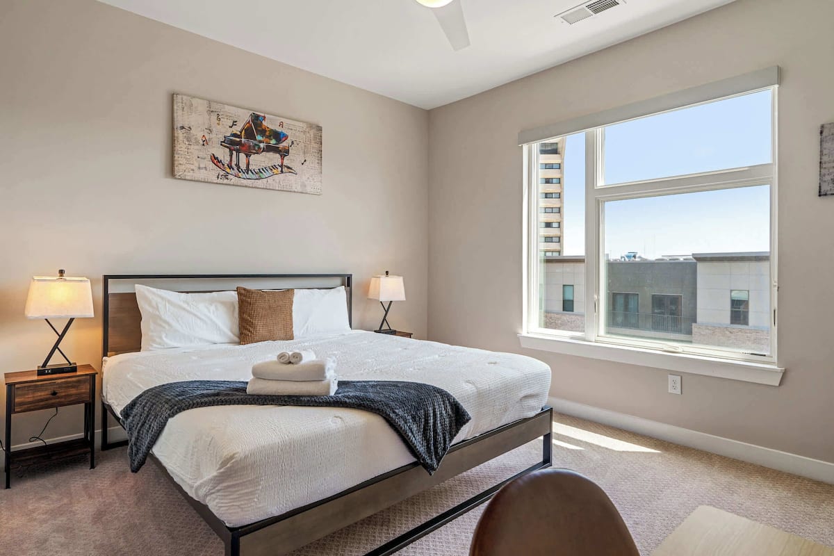 Quarto com uma cama grande, duas luminárias de cabeceira, uma janela com vista para edifícios e um quadro pendurado na parede.
