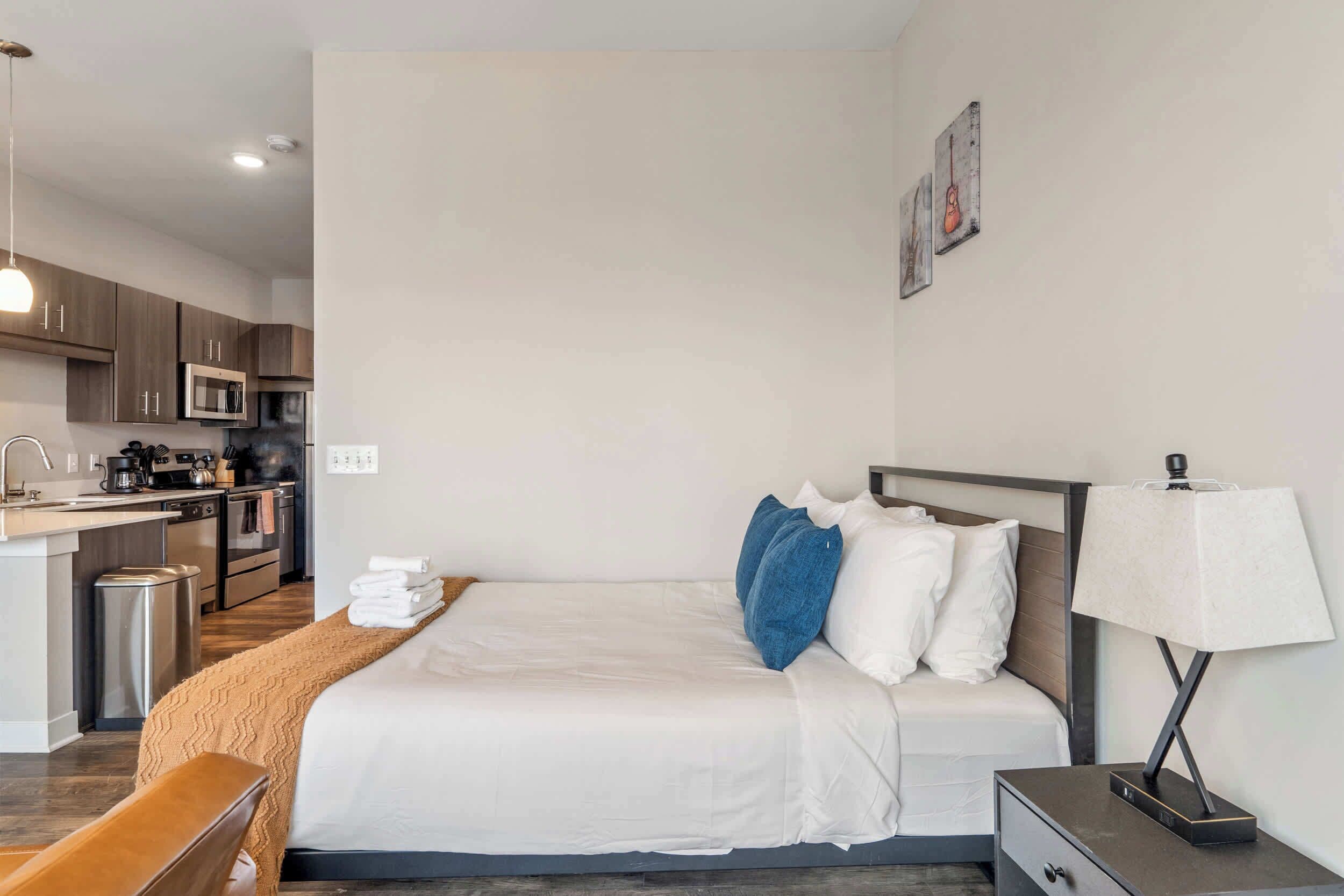 A modern hotel room with a bed, bedside table, lamp, and a kitchenette area.