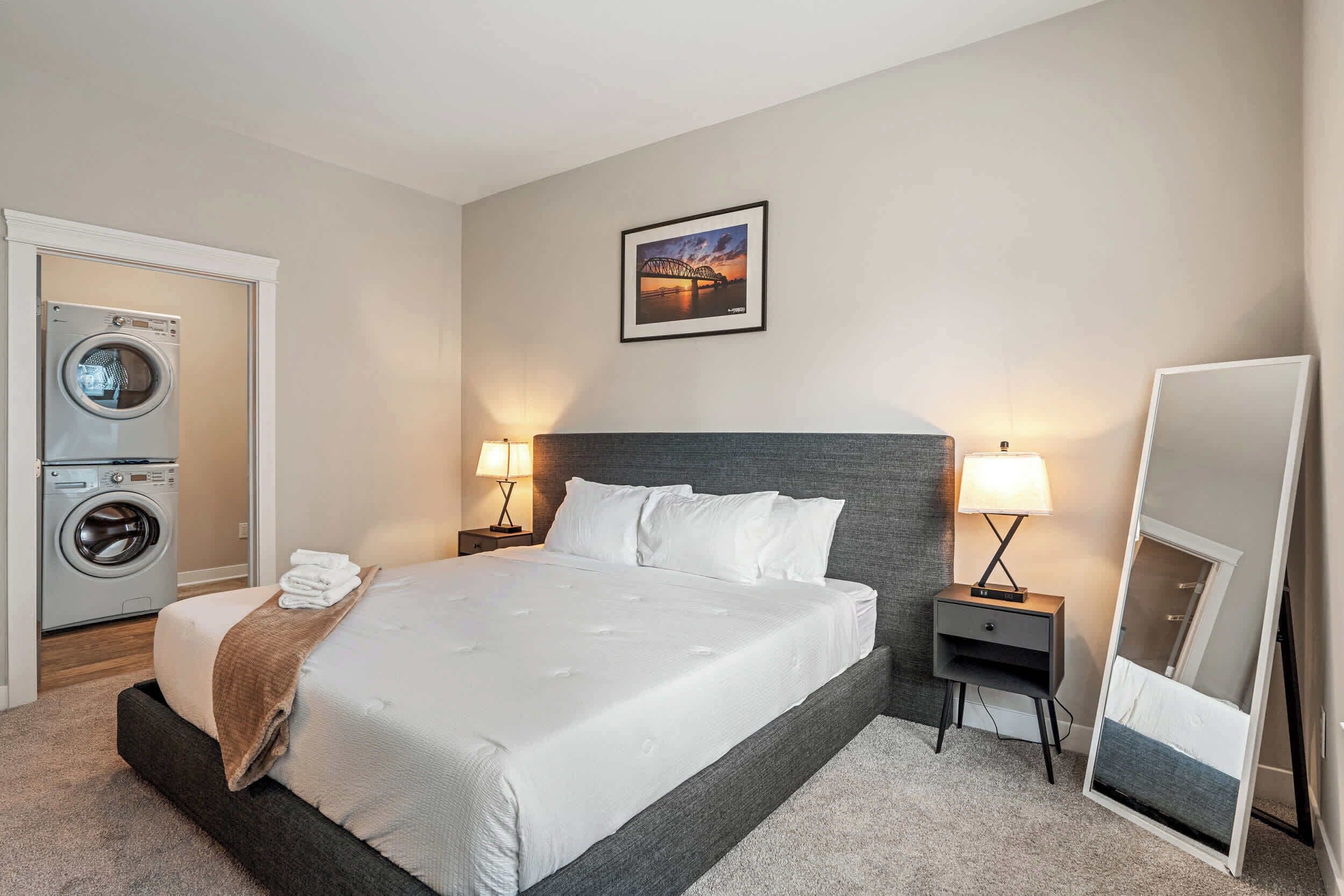 Habitación de hotel con cama, lavadora, mesita de noche con lámpara, espejo y un cuadro en la pared.