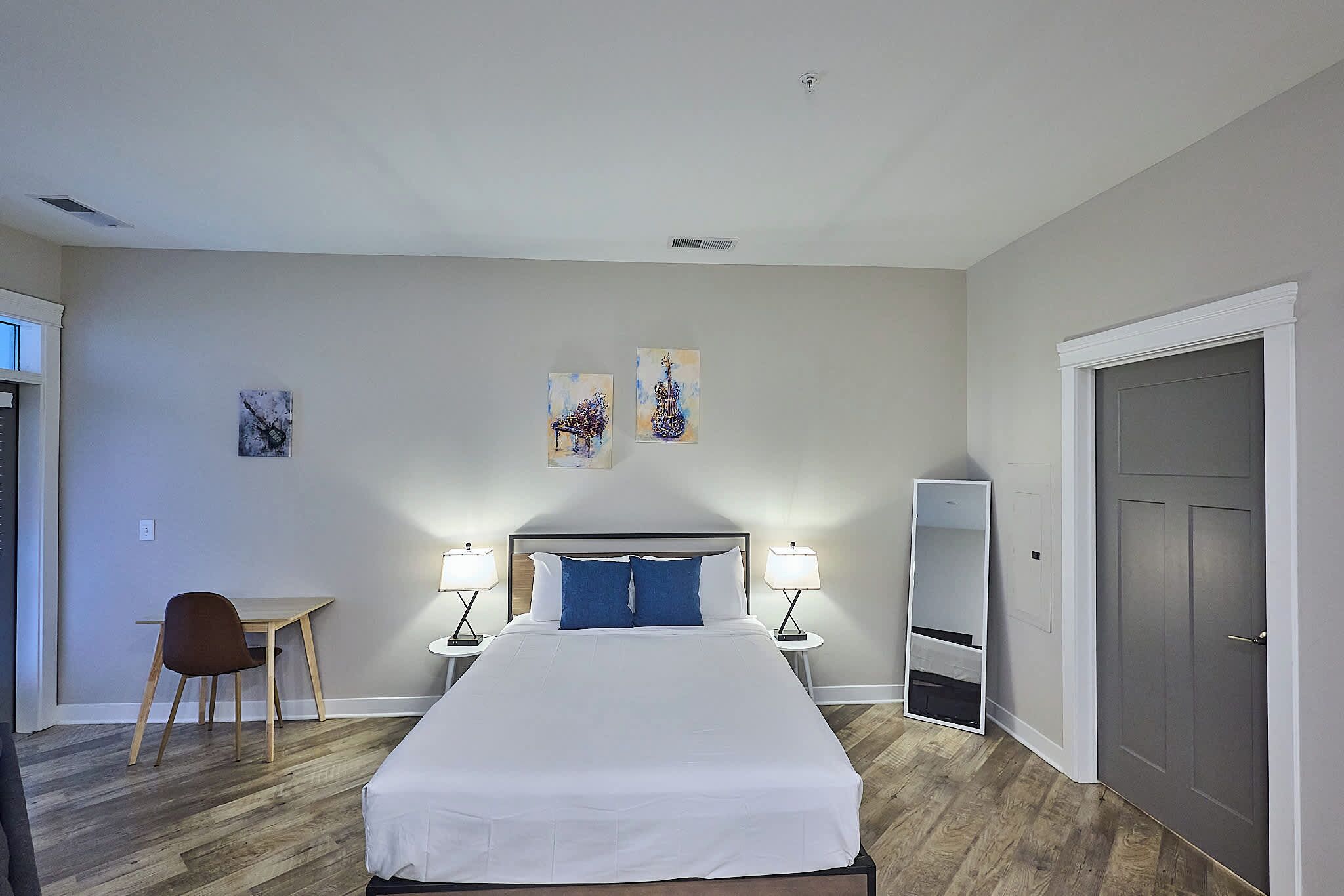 Quarto de hotel com cama, duas luminárias de cabeceira, uma escrivaninha com cadeira e obras de arte na parede.