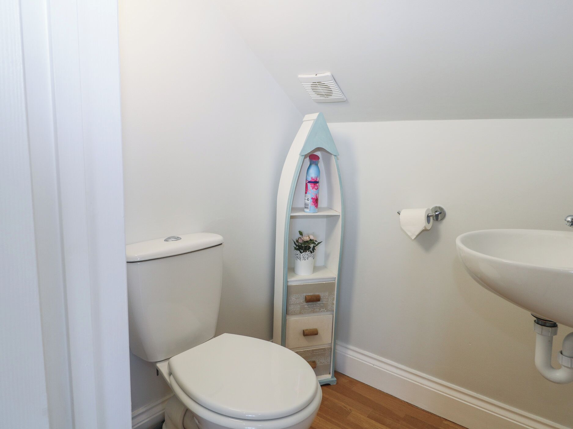 Une petite salle de bain avec des toilettes blanches, un lavabo blanc et une petite étagère sur laquelle se trouvent une bouteille d’eau et une plante en pot.
