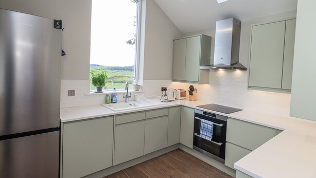 A modern kitchen with a stainless steel refrigerator, a built-in oven, and a window overlooking a green landscape. - Old Cottage (Ulverston)