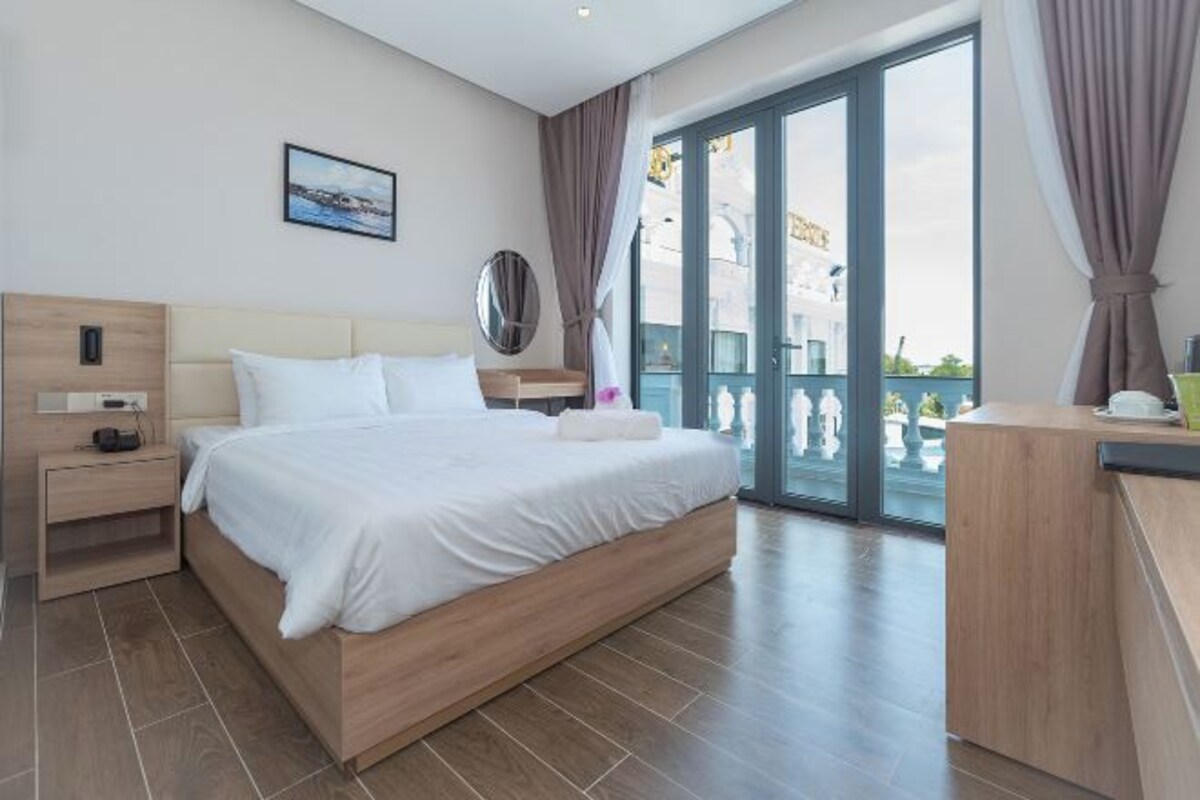 A modern hotel room with a large bed, a wooden nightstand, a desk, and a balcony with a view.